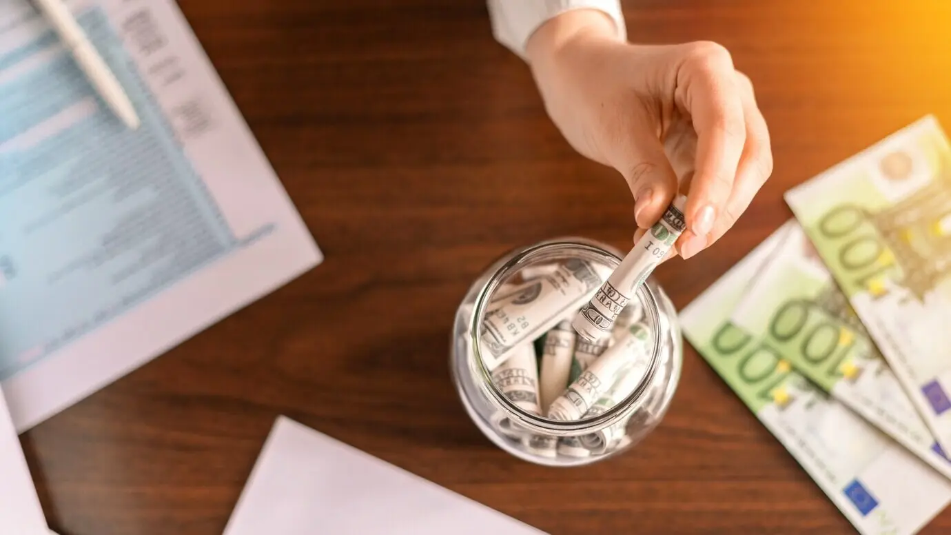 On the table, a woman drops banknotes into a jar containing rolled banknotes. Papers and money are on the table.