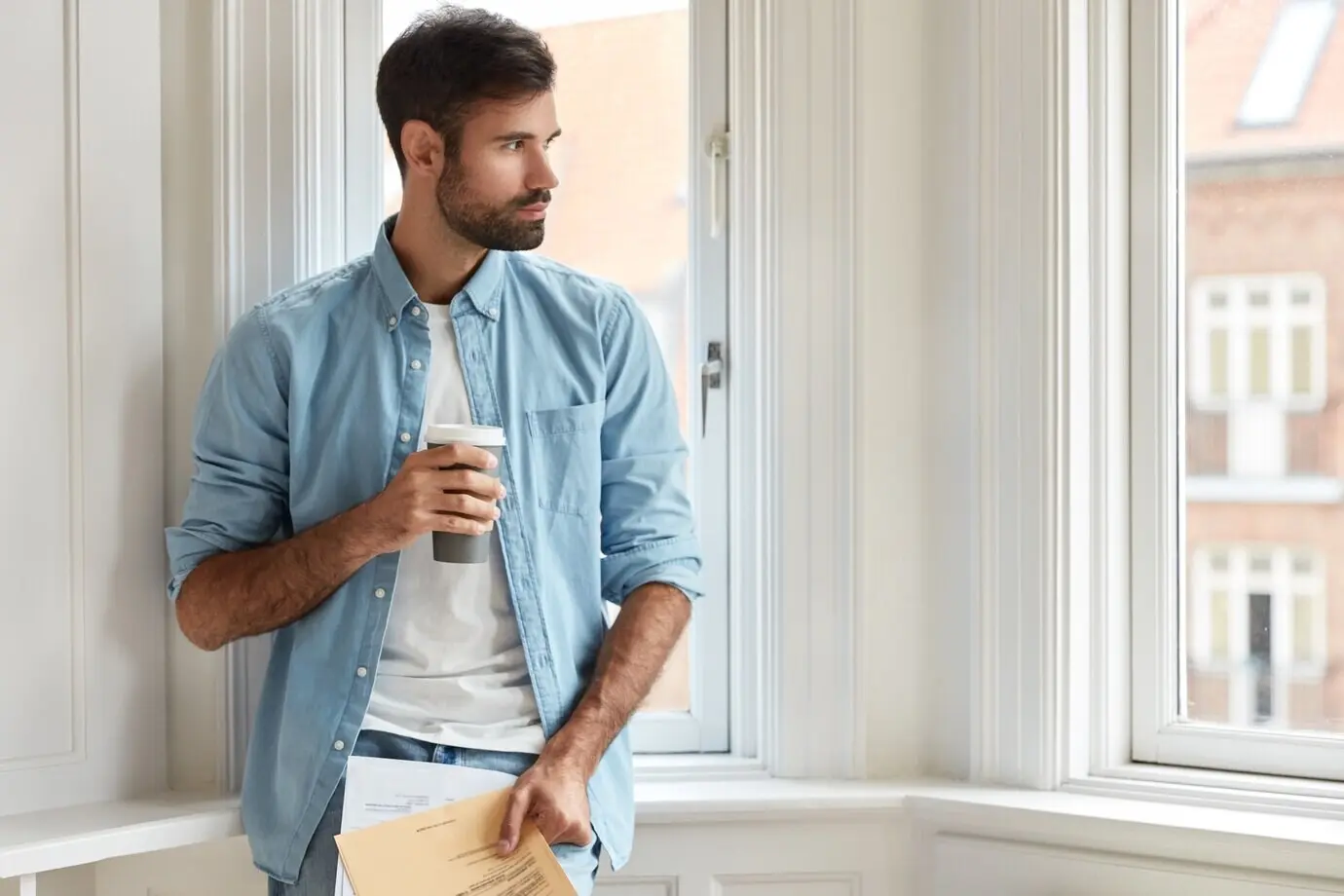 Thoughtful chairman with dark stubble, wearing a stylish shirt, holding a takeaway coffee and papers, reviewing taxes.